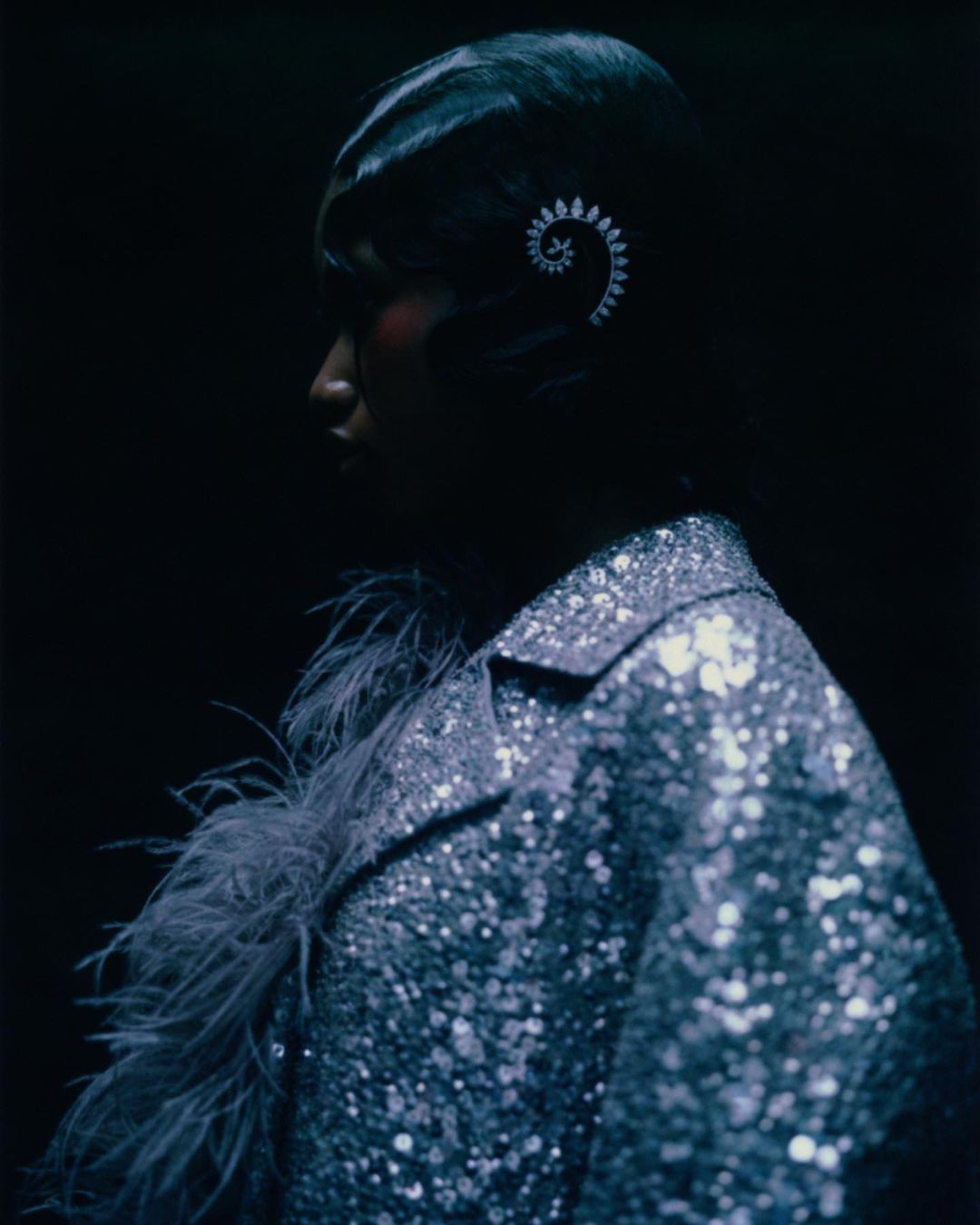 Profile shot of an adult female with short dark hair wearing a sequined jacket with feathers against a dark background.