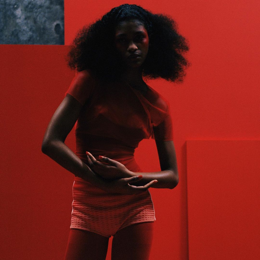 Portrait of a Black woman with voluminous dark hair wearing a red top and shorts against a red studio background by photographer Gabriel Moses.