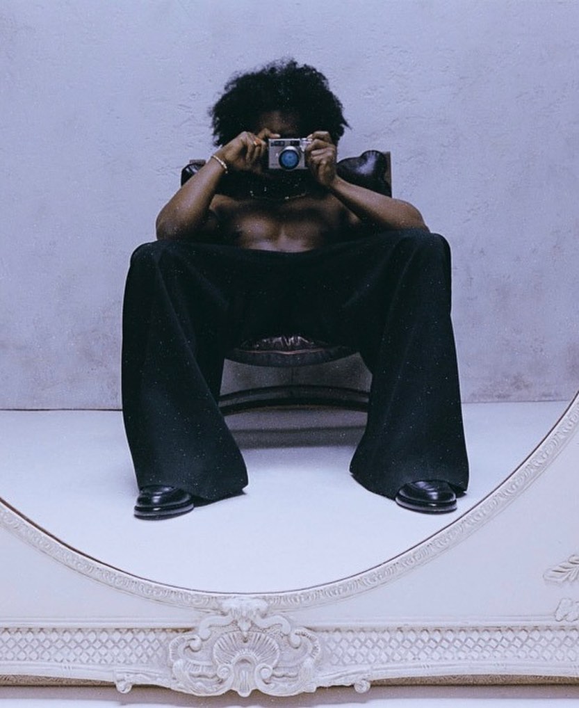 Shirtless man with afro sitting in an armchair holding a camera to his face reflected in an ornate mirror shot by Gabriel Moses