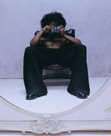 Shirtless man with afro sitting in an armchair holding a camera to his face reflected in an ornate mirror shot by Gabriel Moses