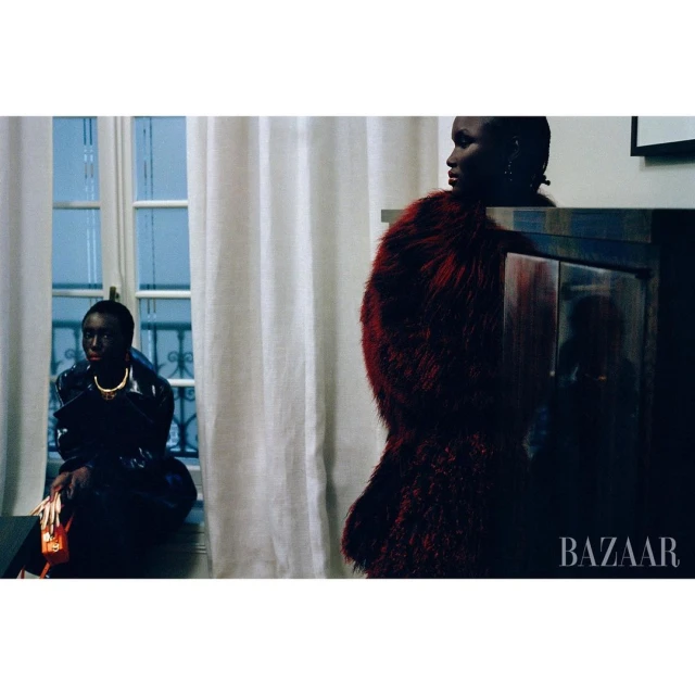Two Black women pose in an indoor setting, one seated near a window, and the other stands near a dark blue cabinet wearing a red fur coat.