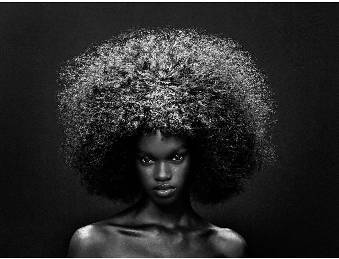 Portrait of a Black woman with a large voluminous afro hairstyle against a dark background by Gabriel Moses