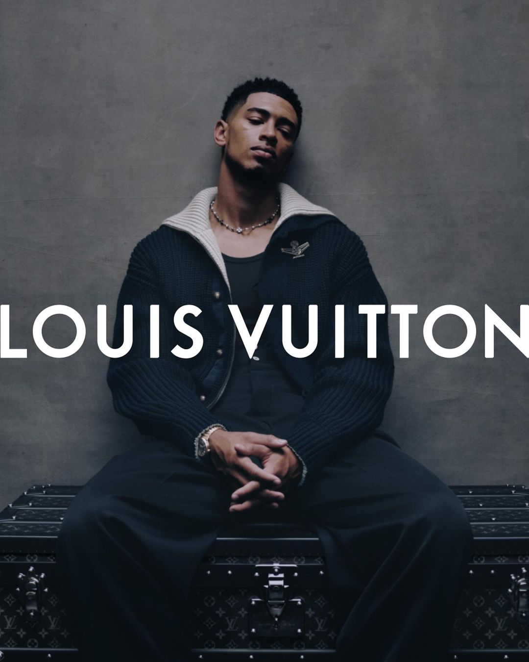 An adult male model sits on a branded Louis Vuitton trunk wearing a black outfit and gold jewelry in a fashion studio shot.