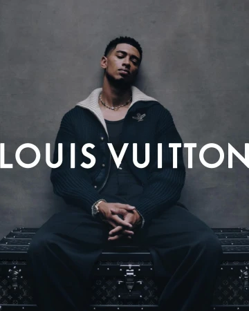 An adult male model sits on a branded Louis Vuitton trunk wearing a black outfit and gold jewelry in a fashion studio shot.