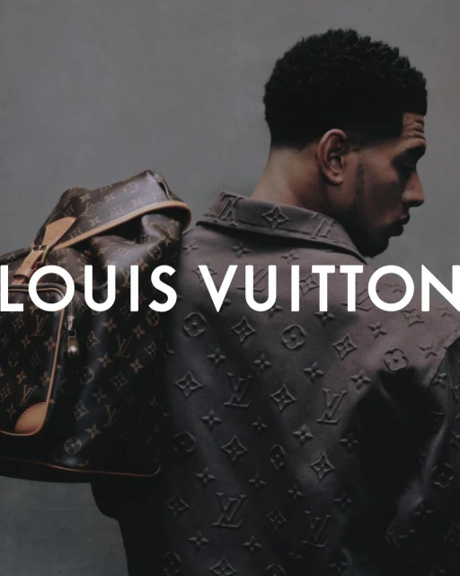 Profile shot of an adult male modeling a Louis Vuitton monogram embossed jacket with a Louis Vuitton duffle bag over his shoulder.