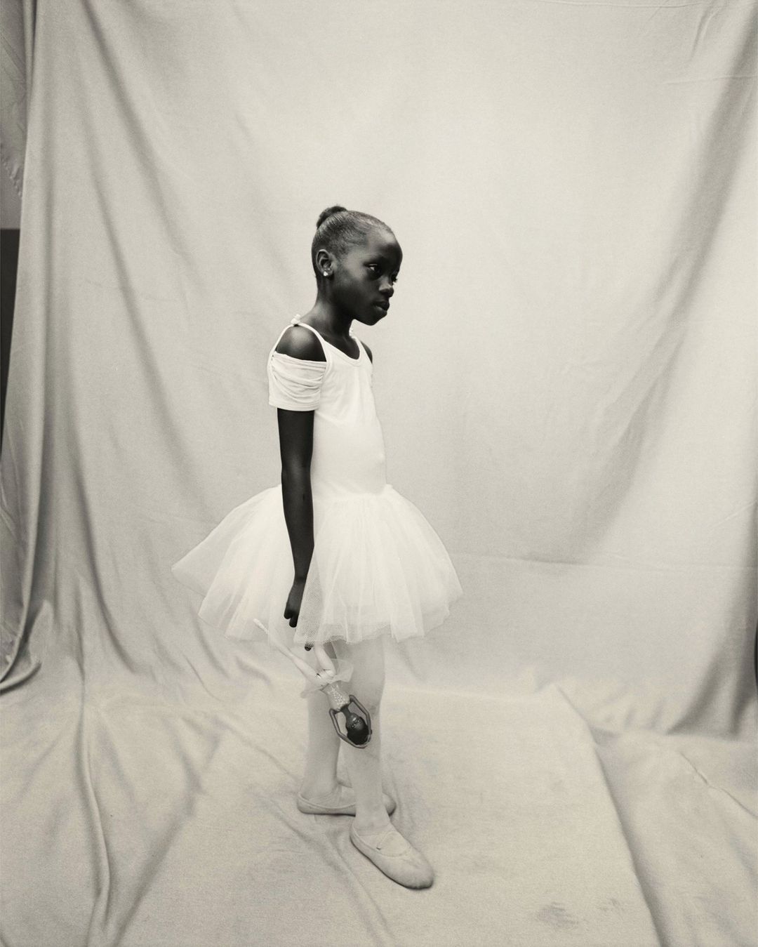 Black and white portrait of a young African American ballerina in a white tutu, holding a doll, against a neutral backdrop.