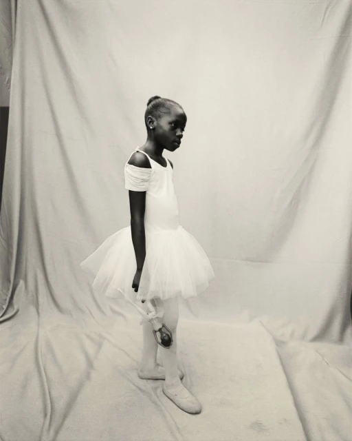 Black and white portrait of a young African American ballerina in a white tutu, holding a doll, against a neutral backdrop.