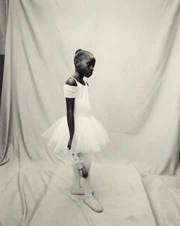 Black and white portrait of a young African American ballerina in a white tutu, holding a doll, against a neutral backdrop.