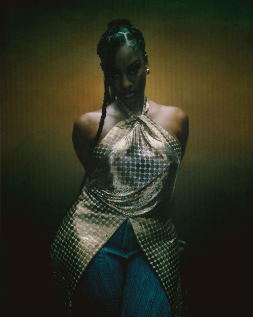 Portrait of a woman in a gold halter top with braided hair against a blurred olive and gold background in a studio setting.