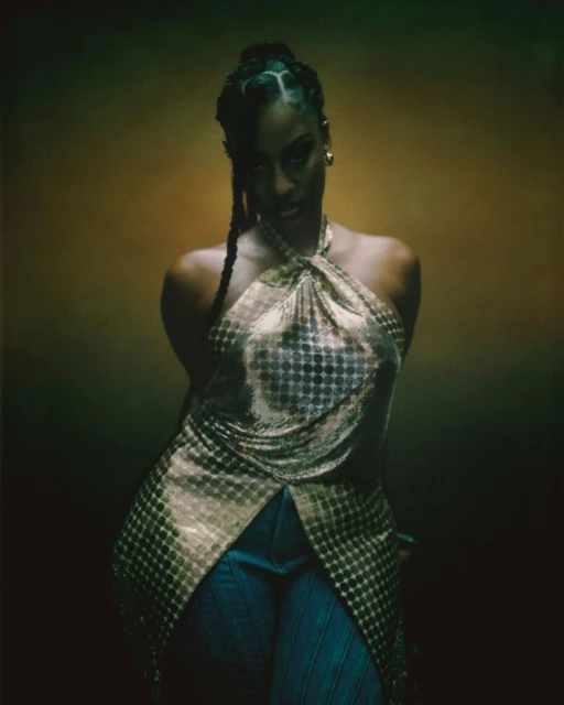 Portrait of a woman in a gold halter top with braided hair against a blurred olive and gold background in a studio setting.