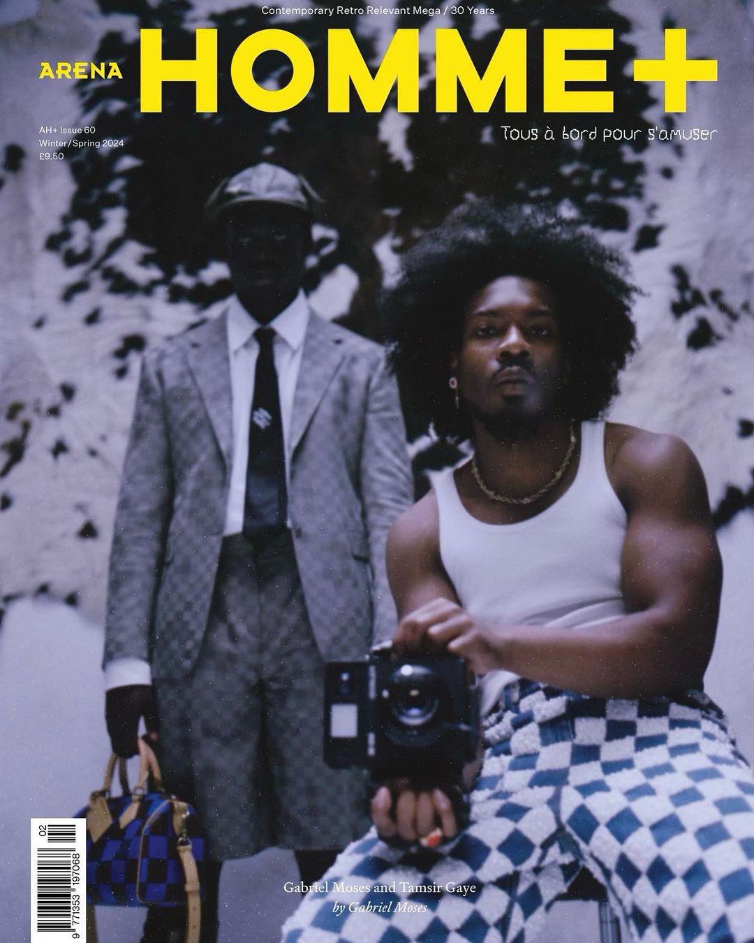 Arena Homme+ magazine cover featuring two men, one holding a camera, in an indoor studio setting with patterned backgrounds and clothing.