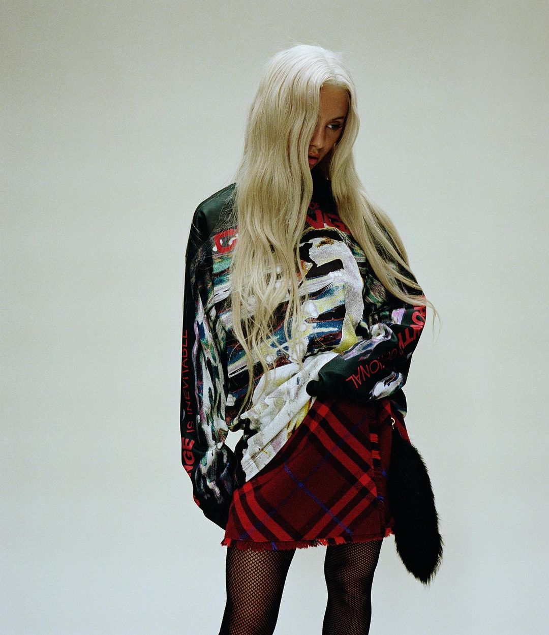 An adult female with long platinum blonde hair poses in a studio setting wearing a graphic top, tartan skirt, fishnet stockings, and black boots.