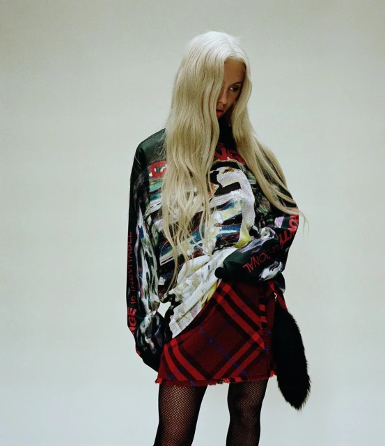 An adult female with long platinum blonde hair poses in a studio setting wearing a graphic top, tartan skirt, fishnet stockings, and black boots.