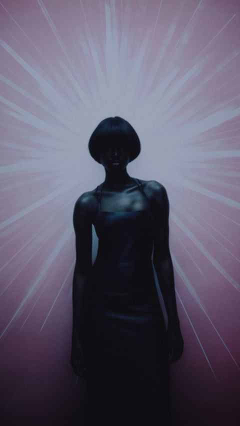 Dark-skinned woman in a black dress stands in front of a pink background with white starbursts, creating a stark and dramatic portrait.