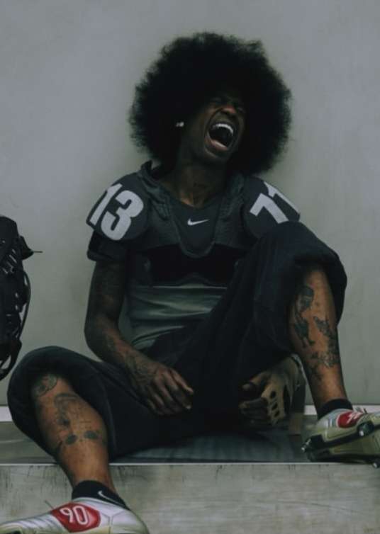 Adult male in athletic gear sits with a large afro and visible tattoos, showing an open mouth expression in front of a plain backdrop.