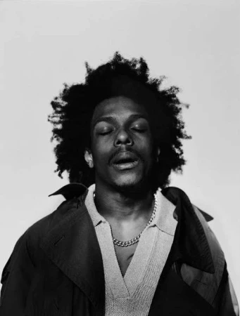 Black and white portrait of a black man with eyes closed wearing a collared shirt, chain necklace, and dark jacket against a plain background.
