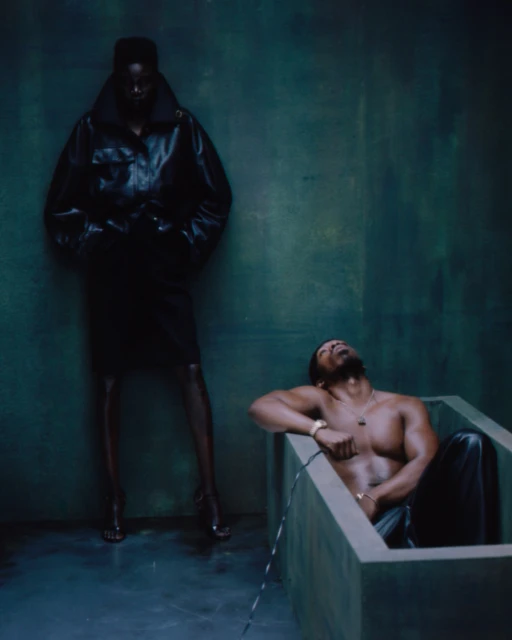 A dark, moody fashion shoot featuring a shirtless man in a tub and a woman in a black leather coat and dress against a green-gray wall.