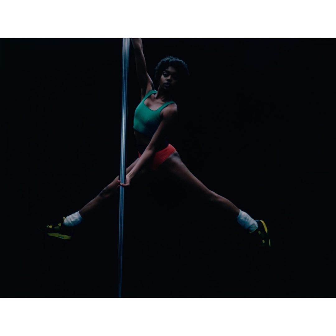 Athletic woman in a green top and red shorts performs a pole dance with legs extended in front of a dark background.