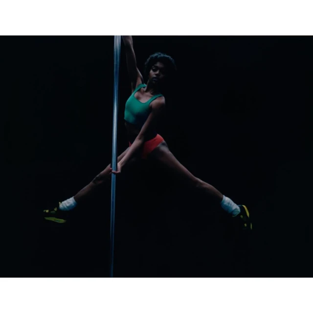 Athletic woman in a green top and red shorts performs a pole dance with legs extended in front of a dark background.