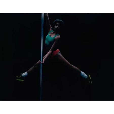 Athletic woman in a green top and red shorts performs a pole dance with legs extended in front of a dark background.