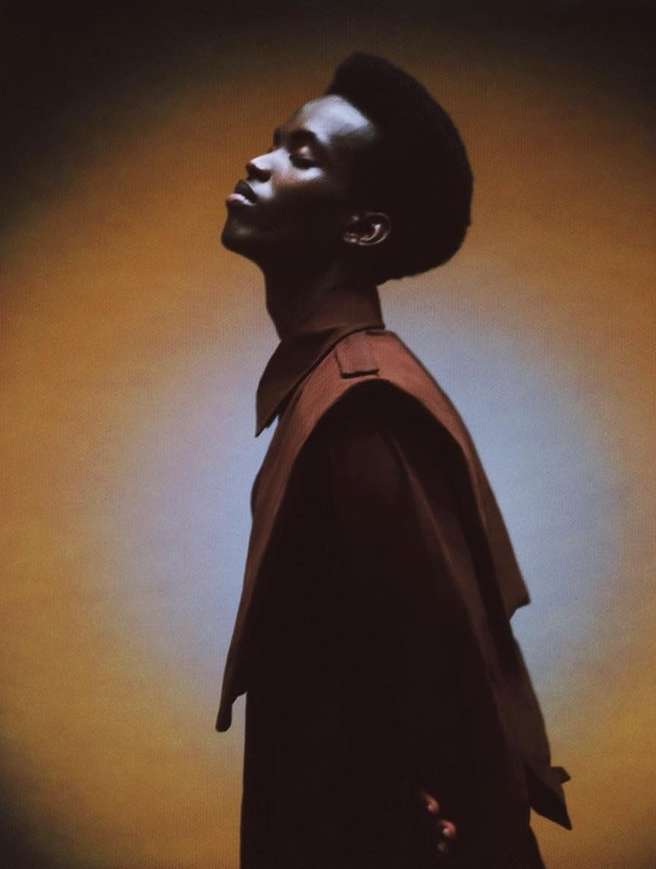 Studio portrait of a stylish young African American man in earth-tone designer clothing against an amber background with dramatic side lighting.