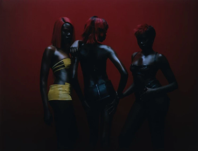 Three women with stylized hair in fashionable outfits pose in front of a red background in a dimly lit studio photograph.