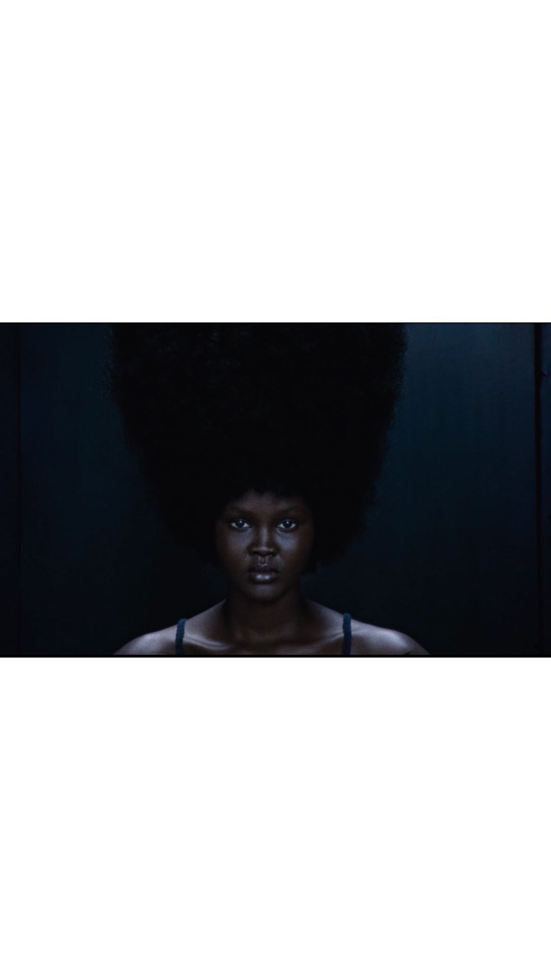 Dark-skinned woman gazes at the camera with a serious expression, large natural hairstyle, and dark backdrop, evoking drama and elegance.