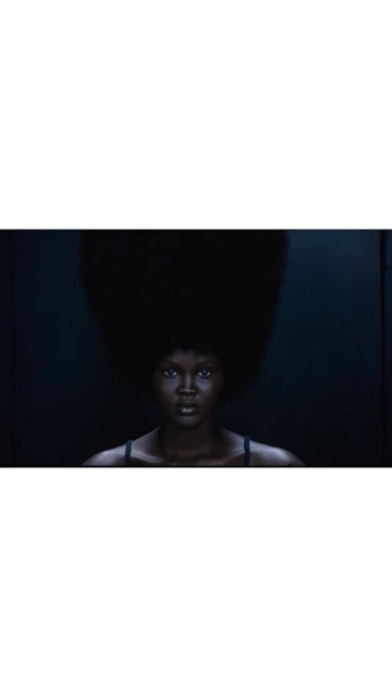 Dark-skinned woman gazes at the camera with a serious expression, large natural hairstyle, and dark backdrop, evoking drama and elegance.