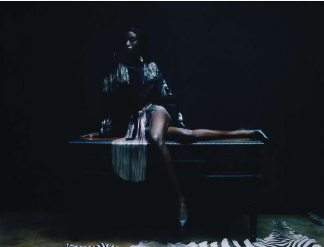 Elegant African American woman in a fringe jacket poses sitting on a chest of drawers in a dark room with zebra-striped rugs.