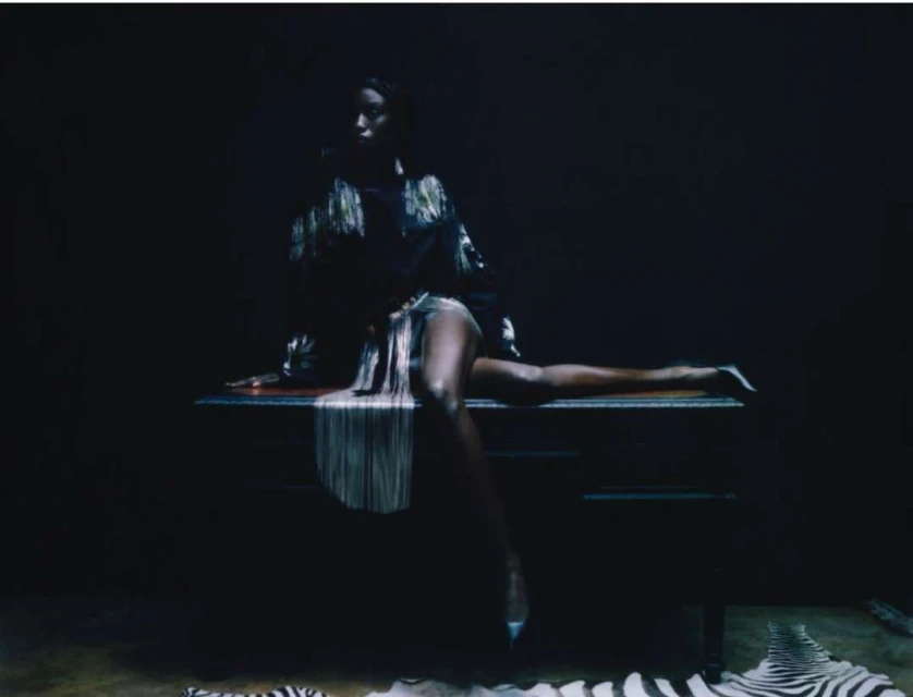 Elegant African American woman in a fringe jacket poses sitting on a chest of drawers in a dark room with zebra-striped rugs.