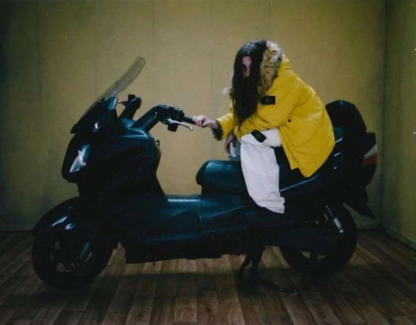 A person with long dark hair, wearing a yellow jacket, is sitting on a black motor scooter indoors on a wooden floor against a yellow wall.