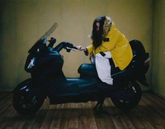 A person with long dark hair, wearing a yellow jacket, is sitting on a black motor scooter indoors on a wooden floor against a yellow wall.