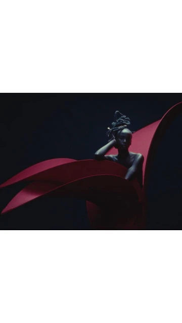 An adult female posing with artistic red fabrics against a dark backdrop in a visually striking beauty or fashion editorial image.