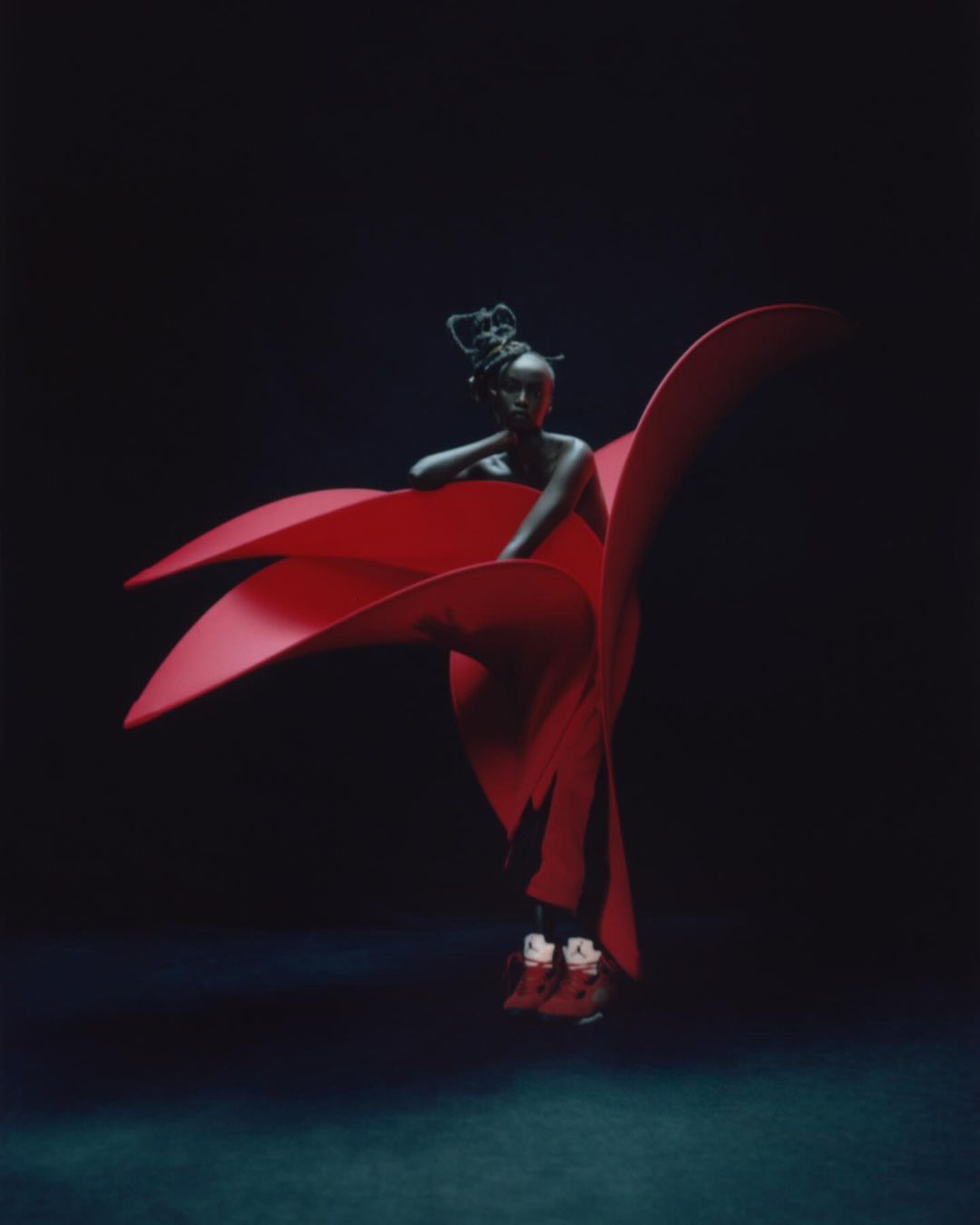 An African American female model in a vibrant red sculptural garment and sneakers against a deep, dark blue backdrop in a studio.