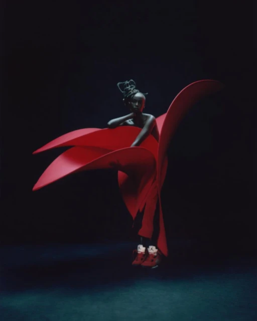 An African American female model in a vibrant red sculptural garment and sneakers against a deep, dark blue backdrop in a studio.