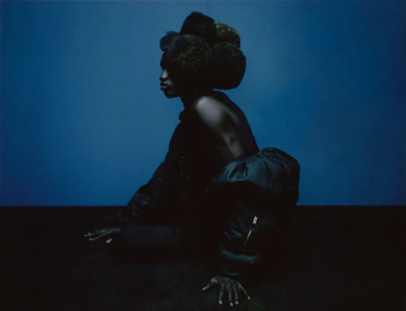 Striking portrait of a Black female model with a dramatic afro hairstyle and dark clothing, posing against a deep blue backdrop in a studio setting.