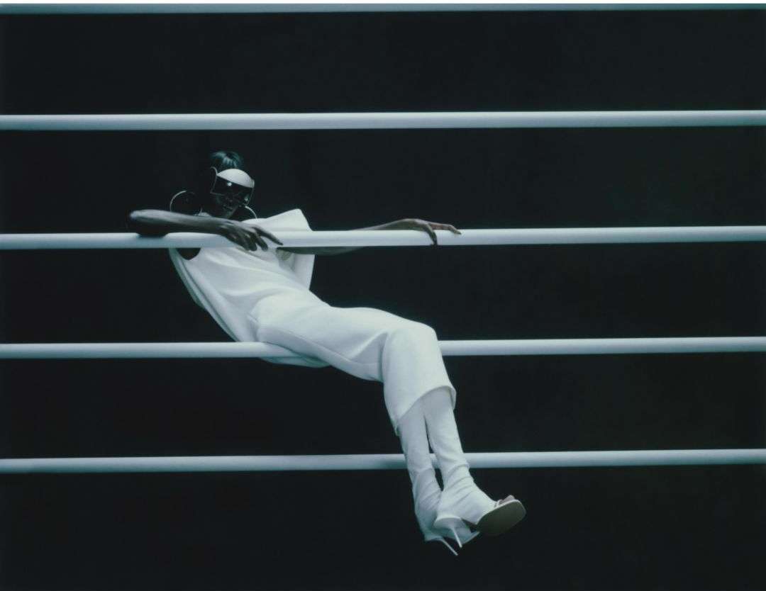 Fashionable adult woman in an all-white outfit poses against horizontal bars in a studio setting, showcasing modern clothing style.
