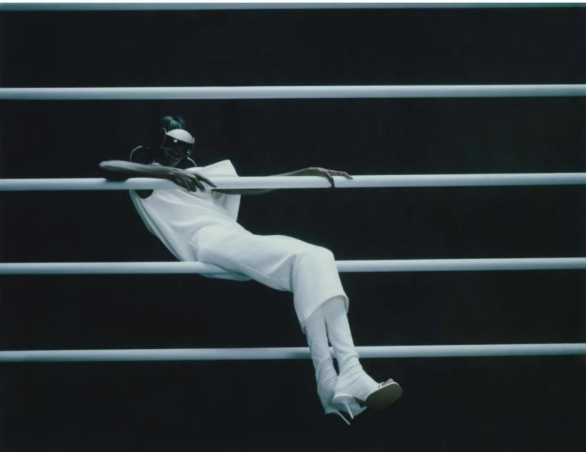 Fashionable adult woman in an all-white outfit poses against horizontal bars in a studio setting, showcasing modern clothing style.