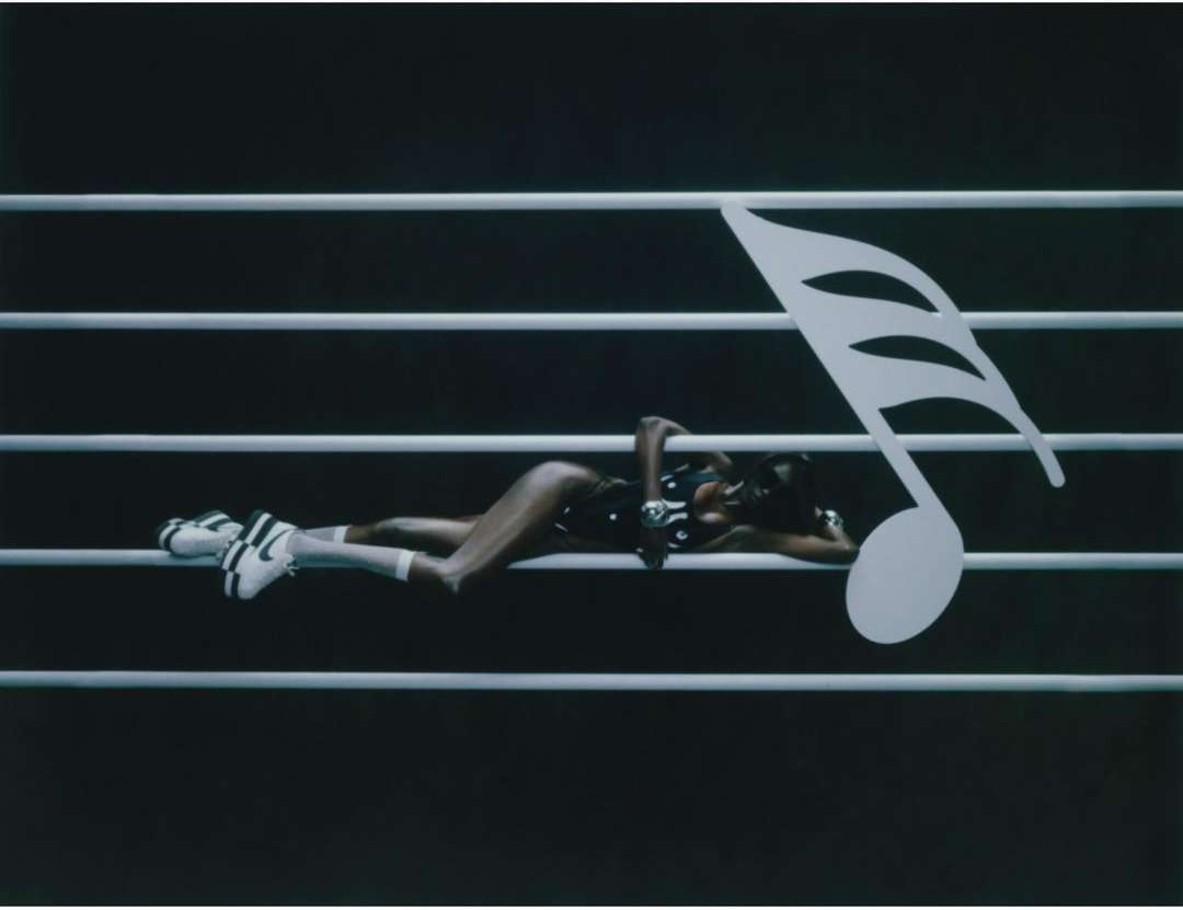 Woman in a bodysuit and sneakers reclines on a musical staff with a large eighth note in this fashion-music crossover scene.
