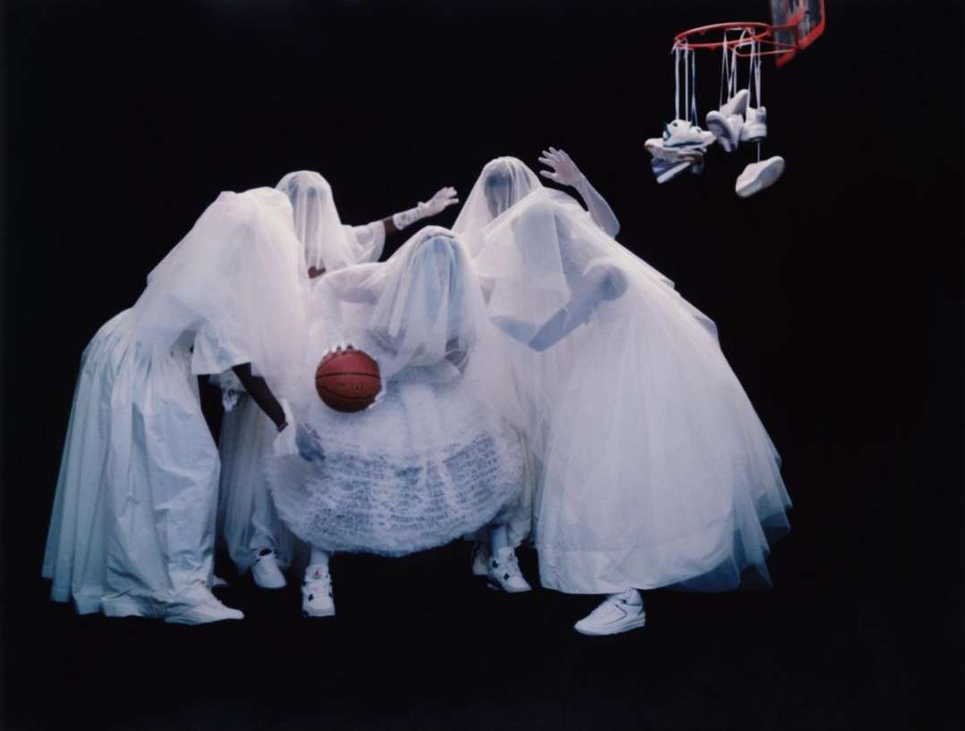 Surreal group of people covered in white fabric around basketball, shoes hanging from hoop against dark backdrop, creating a ghostly effect.