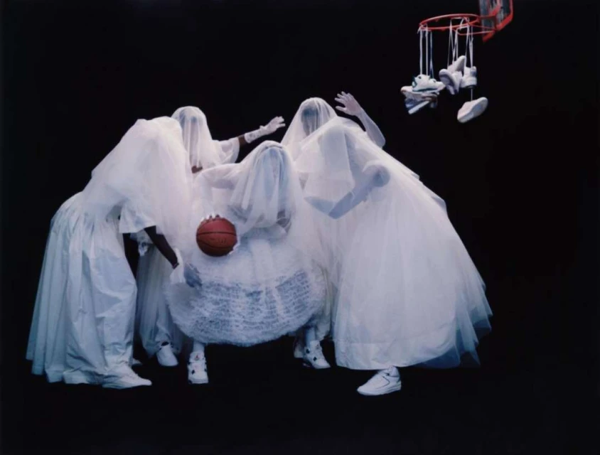 Surreal group of people covered in white fabric around basketball, shoes hanging from hoop against dark backdrop, creating a ghostly effect.