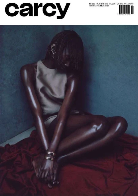 Magazine cover featuring a tanned model in a minimalist tan outfit, seated against a mottled gray background with a red cloth.