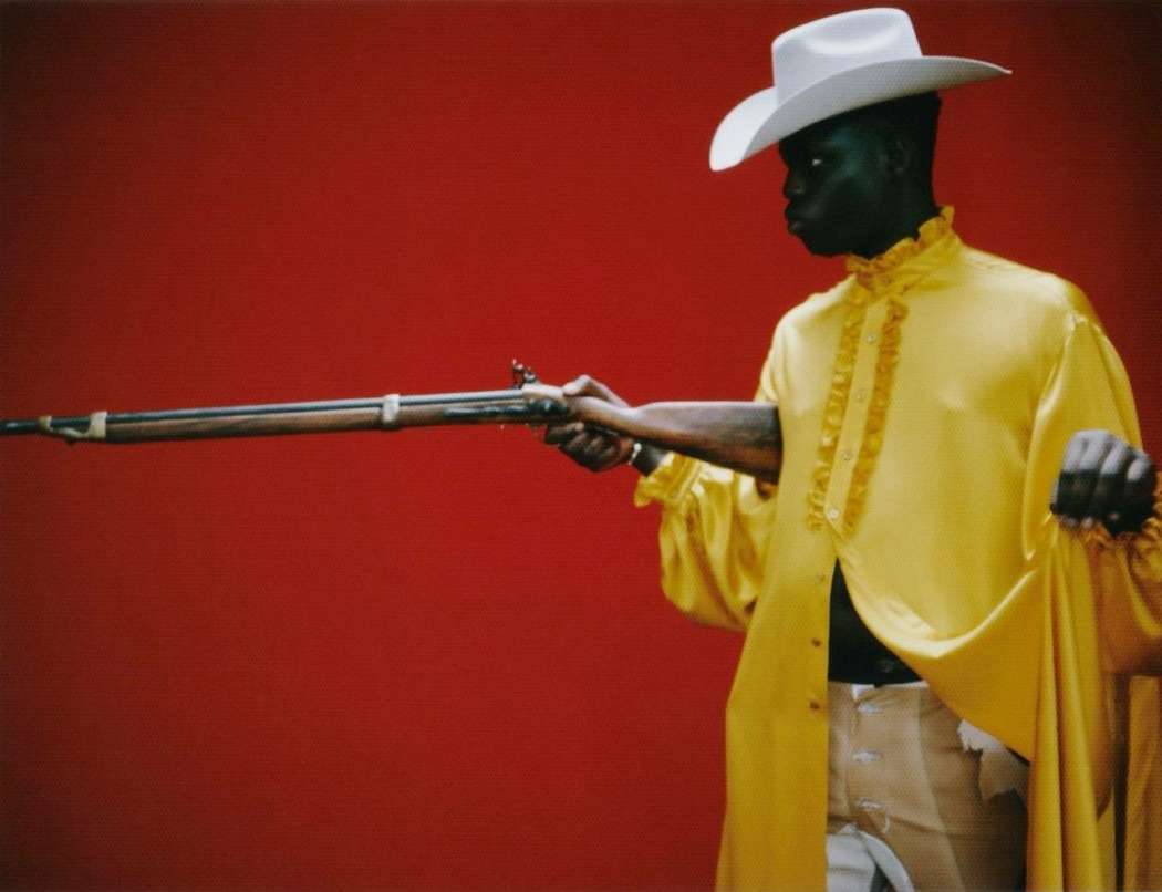 Black man wearing a yellow ruffled shirt and white cowboy hat holding a long rifle against a red background.