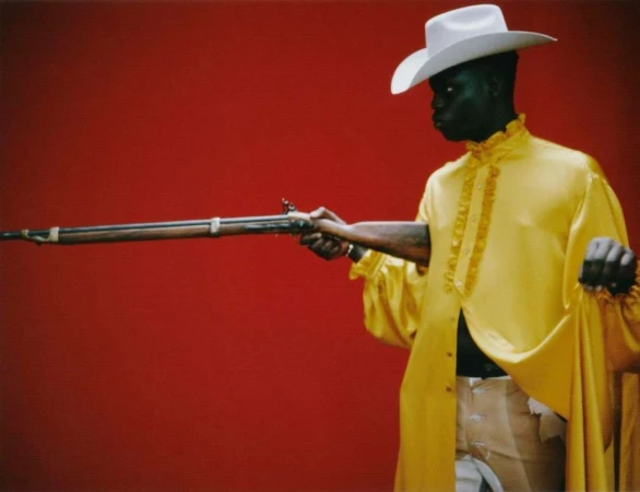 Black man wearing a yellow ruffled shirt and white cowboy hat holding a long rifle against a red background.