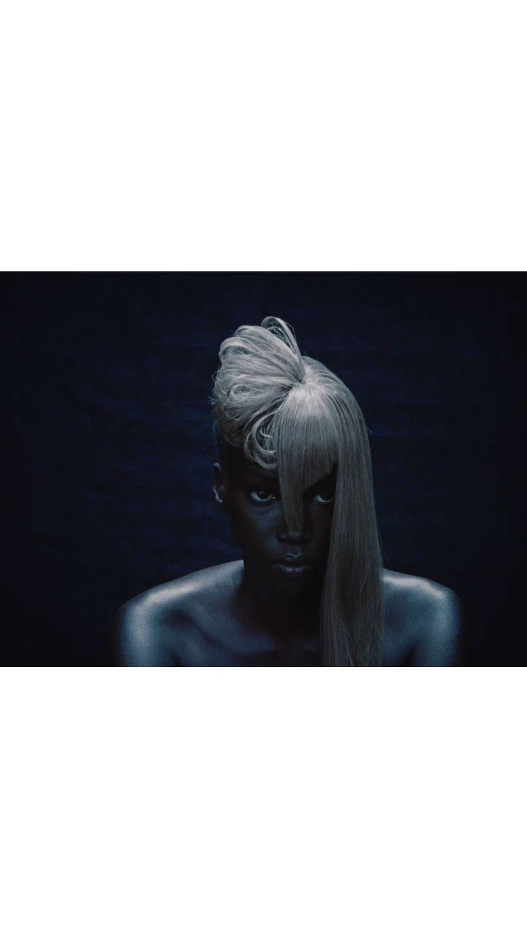 Close-up shot of an adult female with dark skin, a silvery wig with bangs, and dark eye makeup against a dark gray background.