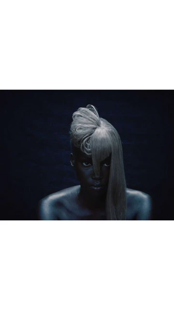 Close-up shot of an adult female with dark skin, a silvery wig with bangs, and dark eye makeup against a dark gray background.