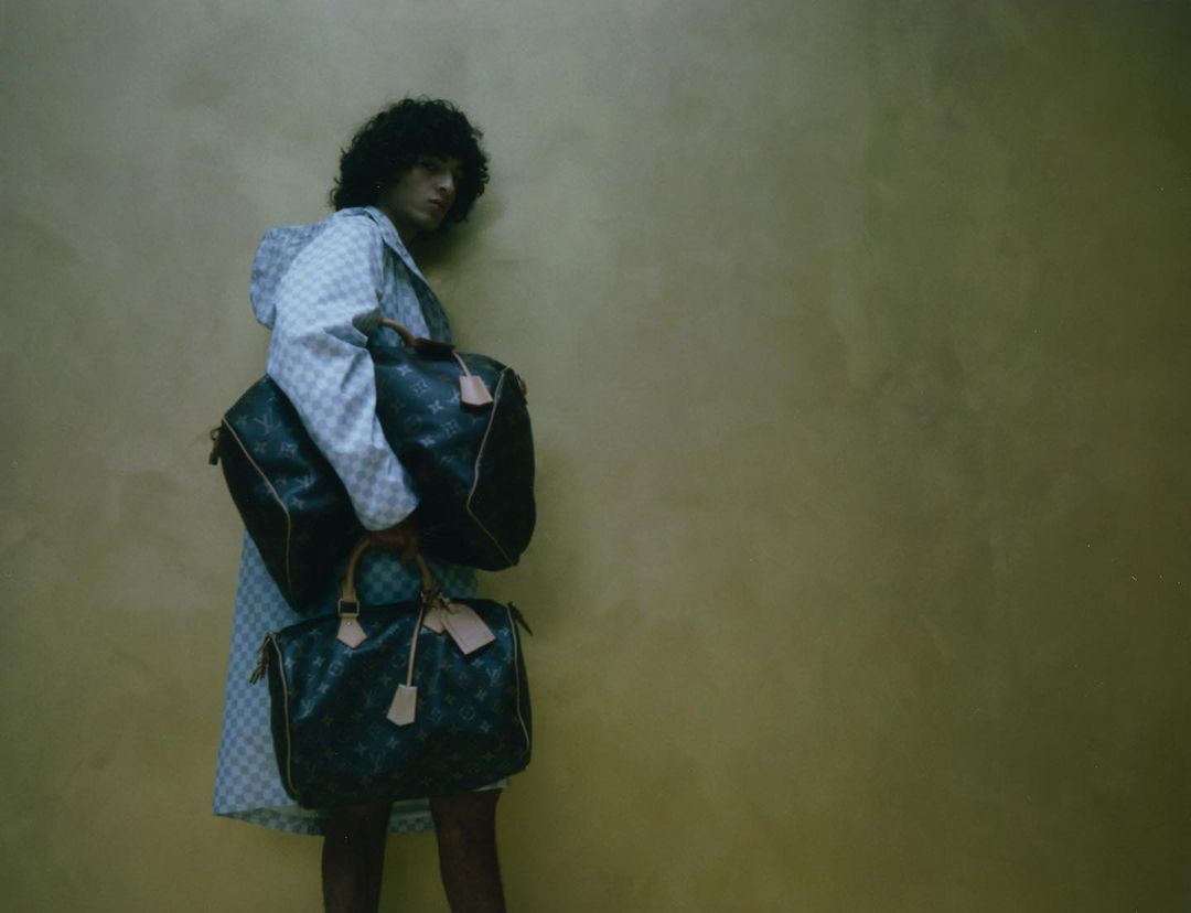 Person with curly hair and checkered jacket holding Louis Vuitton duffel bags against mottled brown backdrop in eye-level medium shot.
