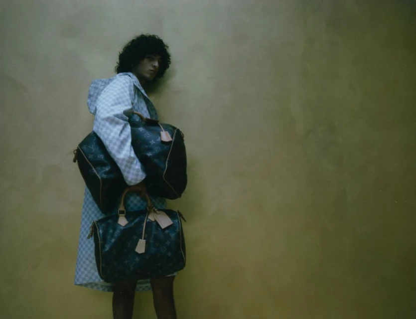 Person with curly hair and checkered jacket holding Louis Vuitton duffel bags against mottled brown backdrop in eye-level medium shot.