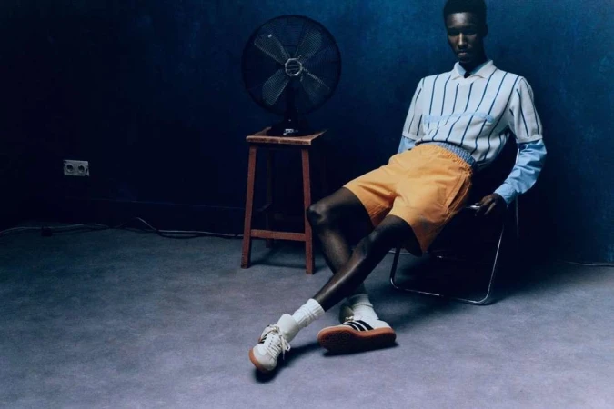 A dark-skinned man in shorts and a striped shirt reclines on a chair in a minimalist space, featuring a fan, stool, and moody lighting.