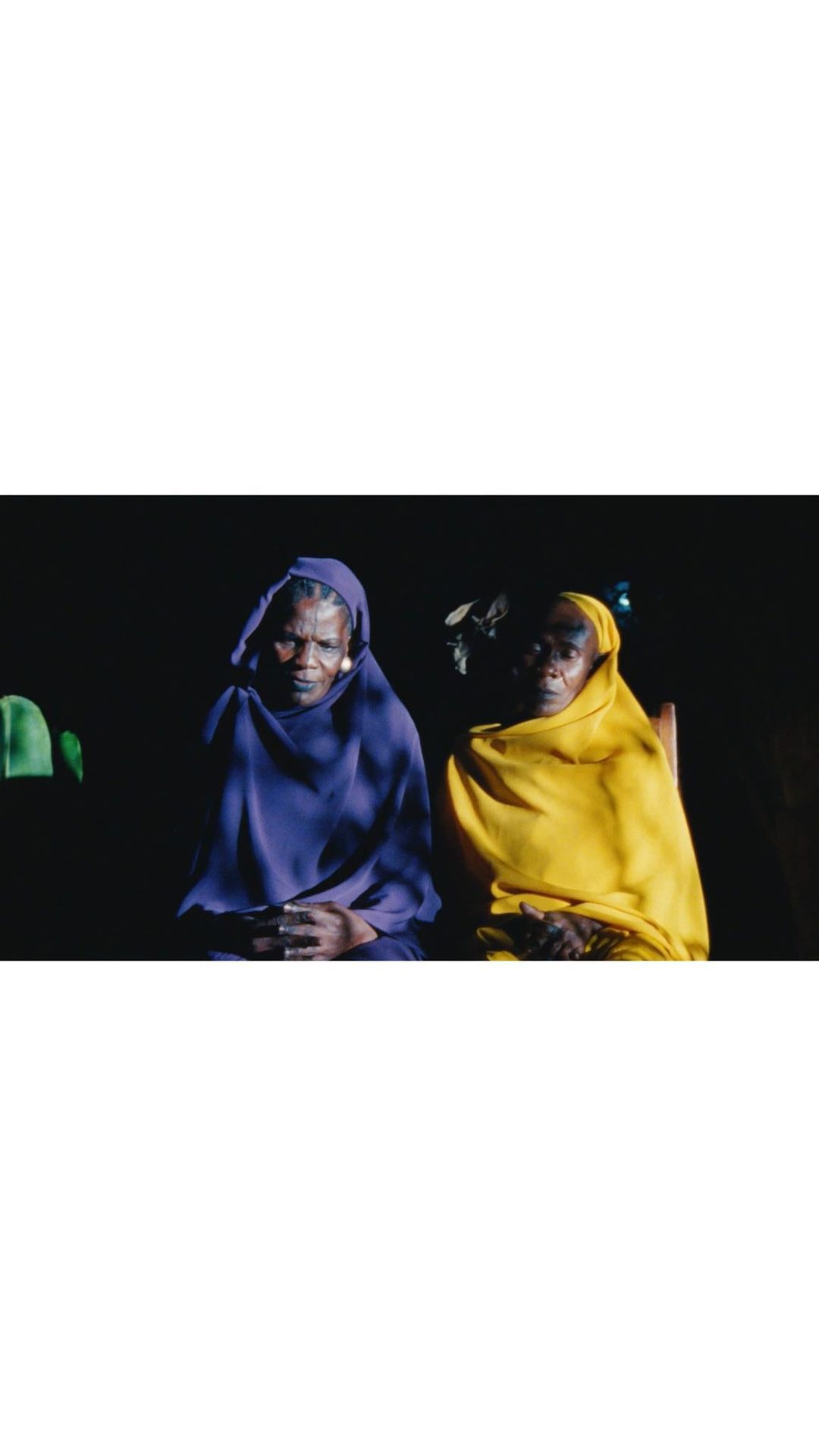 Two elderly African women sit indoors wearing colorful headscarves and robes, their hands clasped in their laps in a dark room.